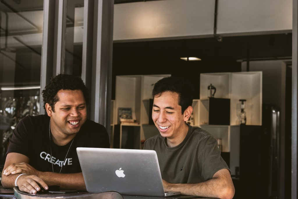An image of two smiling mean sitting at a desk looking at a laptop