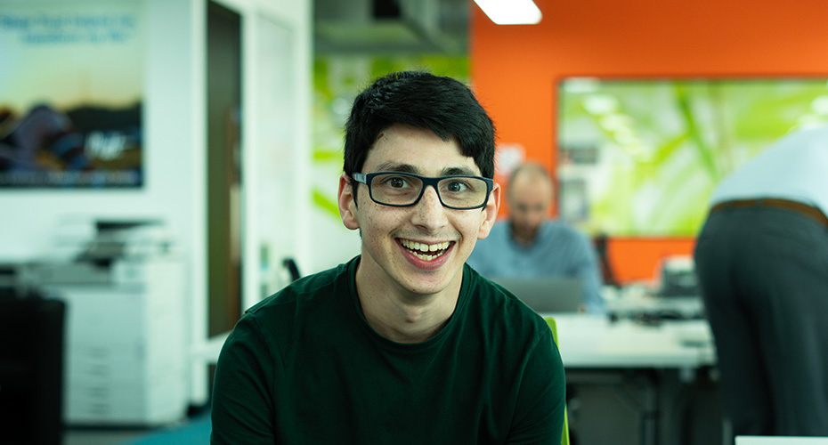 An image of a young man, seated in an office. He smiles and wears glasses.