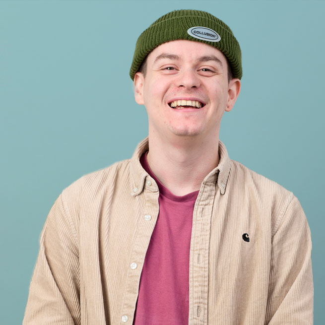 An image of a man smiling, wearing a small beanie hat, wearing a shirt over a t-shirt