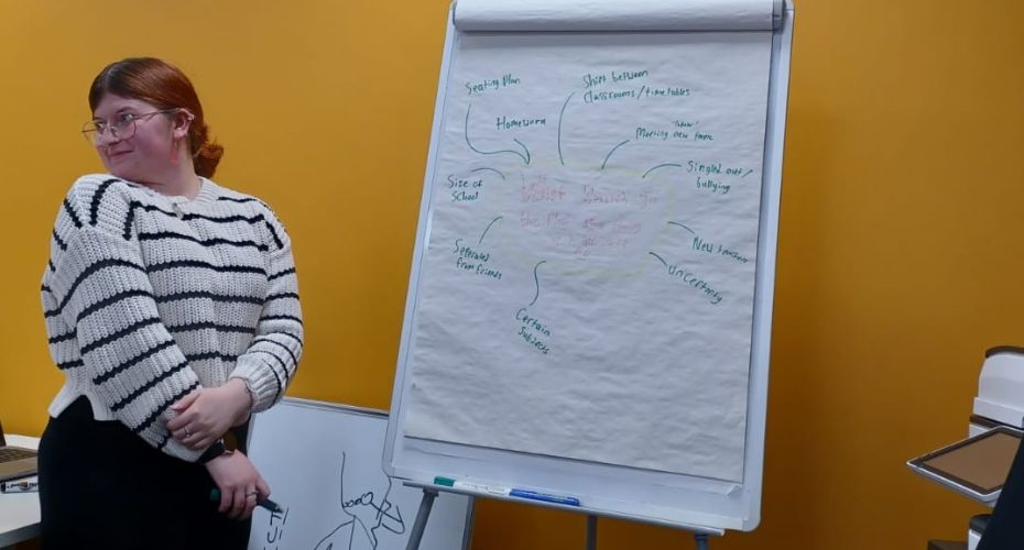 Leah wearing glasses and a white and black jumper stood next to a flip chart making notes