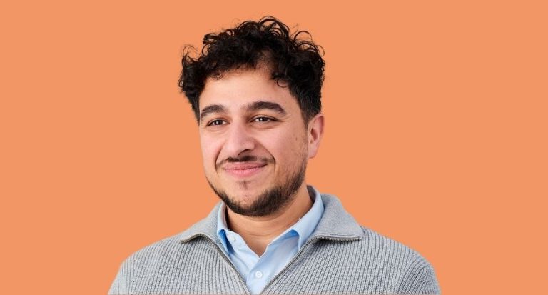 An orange background with a man, from shoulders upwards smiling. He has short dark hair with a thin moustache and beard. He is wearing a pale blue shirt with a grey ribbed jumper over the top. Text above him reads “Over 1,000 people have found, returned or remained in employment with Working Win.”