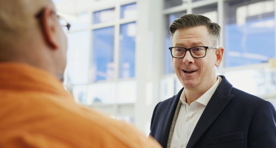 Photo of a man wearing a black suit jacket and white shirt talking to another person.