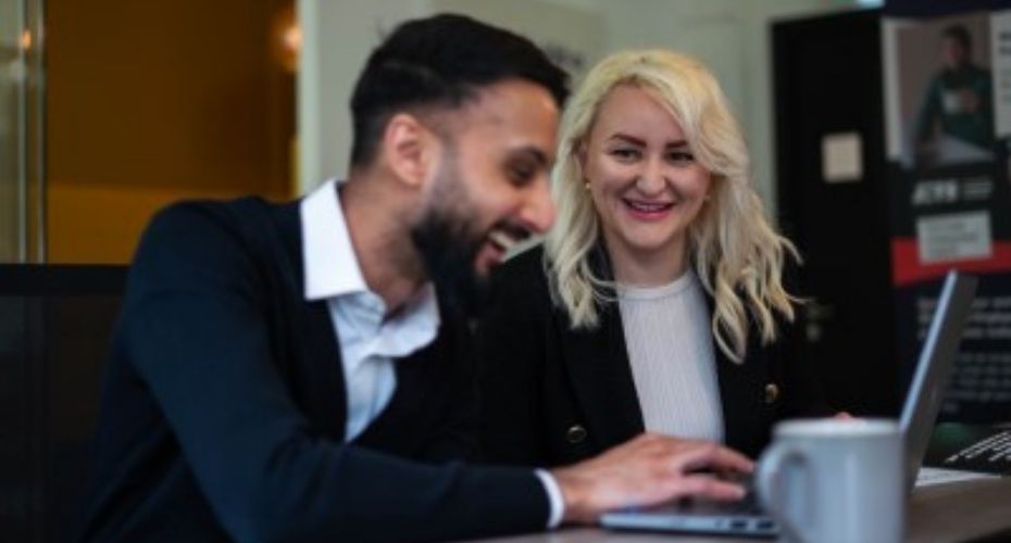 Man and woman smiling and talking while sitting at a desk and working on a laptop.