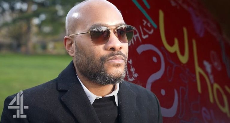 Photo of a man wearing sunglasses stood in front of a wall with writing on it.