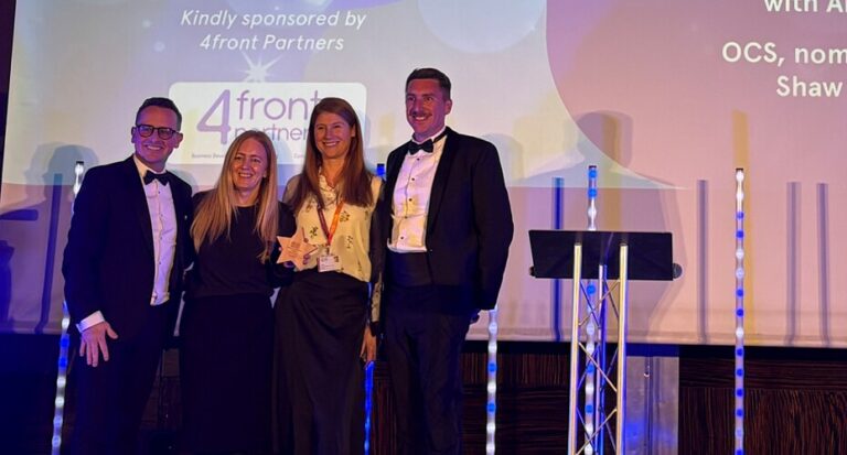 Image shows four people standing on the ERSA stage smiling. They are dressed smartly and at the back of the stage, the finalist's names are projected onto a screen.