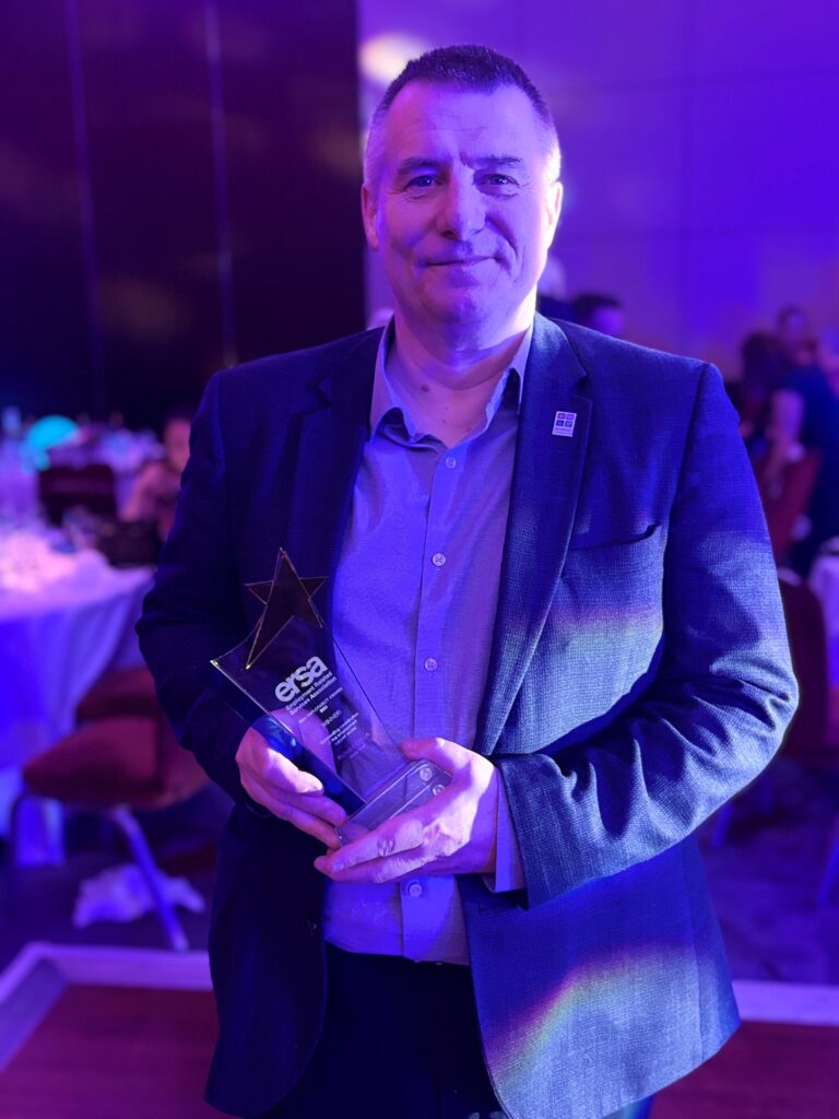 Image of CCO, Richard Clifton, standing in a smart suit and holding his trophy with a smile.