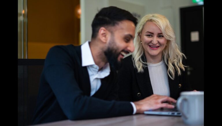 A man and a woman sit talking and smiling in front of a laptop.